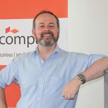 L'image représente un homme debout devant un mur blanc et orange, avec une montre au poignet gauche et une chemise à boutons bleu clair.