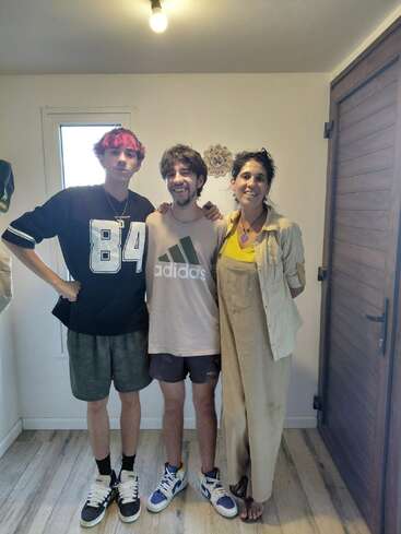 Three people pose together in a room. The person on the left has pink hair, wearing a black shirt. Everyone is smiling, standing relaxed on a wooden floor.