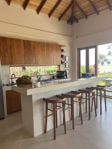 Esta cozinha moderna tem um teto alto de madeira, armários elegantes, uma ilha de concreto com quatro bancos de madeira, luz natural e vista para a vegetação lá fora.