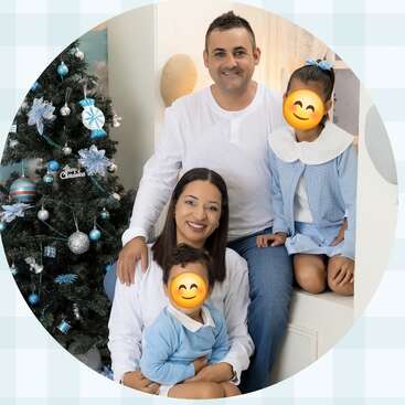 A family of four poses happily beside a decorated Christmas tree. The parents smile while the children’s faces are covered with smiley emojis. Cozy, festive atmosphere.