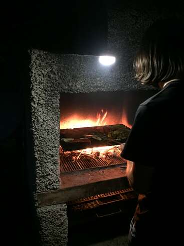 La imagen muestra a una persona de pie delante de una parrilla, con un fuego ardiendo en su interior, sobre un fondo oscuro.