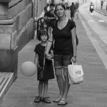 The image depicts a woman and a child standing on a sidewalk, holding hands, with the woman carrying shopping bags and the child holding a balloon.