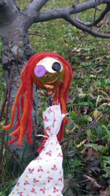 The image depicts a whimsical outdoor scene featuring a handmade doll with vibrant red hair and a floral dress, situated in front of a tree trunk.