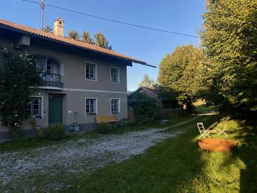 La imagen muestra una casa de dos plantas con exterior marrón, tejado de tejas y chimenea, rodeada de árboles y un patio con césped y entrada de grava.