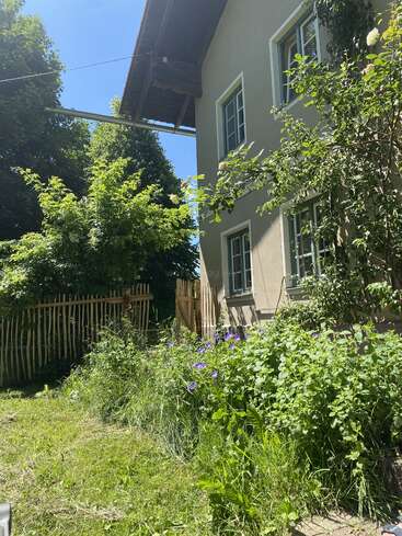 La imagen representa el lateral de una casa con jardín, con una valla, árboles y arbustos, sobre un cielo azul despejado en un día soleado.
