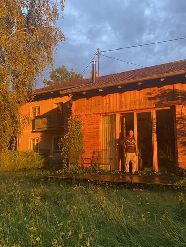 La imagen representa una escena serena de un hombre en el porche de una casa rústica, rodeado de exuberante vegetación y un cielo nublado.