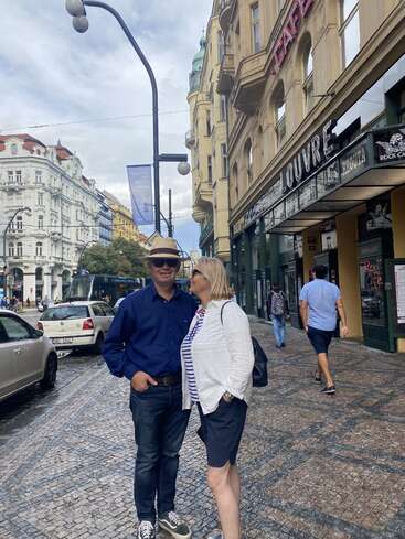 La imagen muestra a un hombre y una mujer de pie en una calle adoquinada, rodeados de edificios y vehículos, con la cabeza de la mujer vuelta hacia el hombre.
