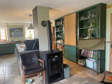 Esta acogedora habitación cuenta con una estufa de leña, estanterías verdes con platos y libros, una isla de cocina, un sillón, suelo de baldosas y grandes ventanas que aportan luz natural.