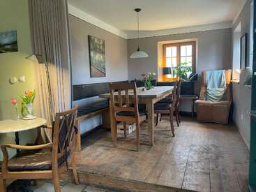 Acogedora zona de comedor con mesa de madera, sillas y bancos. El sillón junto a la ventana, las plantas, la lámpara colgante, las flores y la luz del sol crean un ambiente cálido.