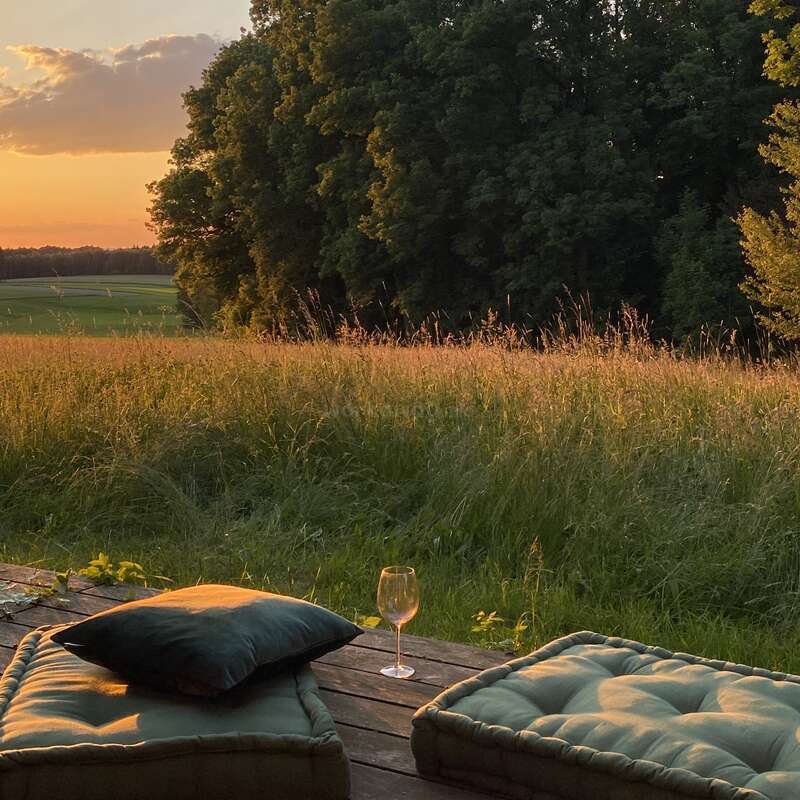 La imagen representa un entorno sereno al aire libre con dos cojines en una terraza de madera, una copa de vino y una vista pintoresca de un campo y árboles al atardecer.
