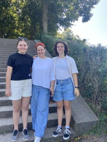 Three women stand smiling on outdoor stone steps surrounded by greenery. They wear casual summer clothes: shorts, t-shirts, and sunglasses. It’s a bright, sunny day.