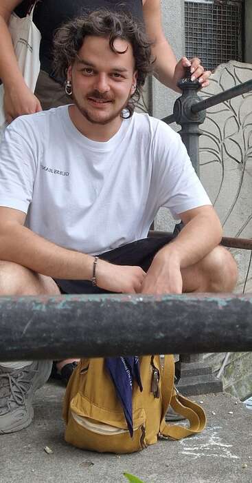 Ein junger Mann mit lockigem Haar und einem weißen T-Shirt hockt lächelnd auf einer Treppe. Vor sich hat er einen gelben Rucksack, und er trägt Freizeitkleidung.