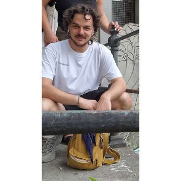 A young man with curly hair and a white T-shirt crouches on steps, smiling. He has a yellow backpack in front of him and casual attire.