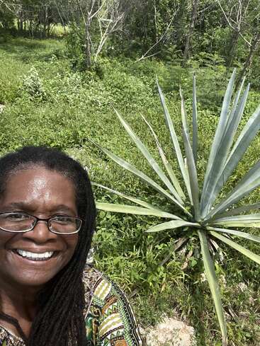 A smiling woman with glasses and long hair takes a selfie outdoors beside a large agave plant, surrounded by lush, green vegetation and trees.