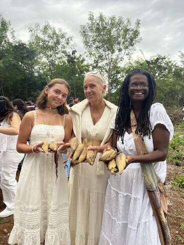 Drei weiß gekleidete Frauen stehen im Freien, lächeln und halten Mais in der Hand. Üppig grüne Bäume umgeben sie, während sich im Hintergrund eine kleine Menschenmenge versammelt und eine fröhliche Atmosphäre schafft.