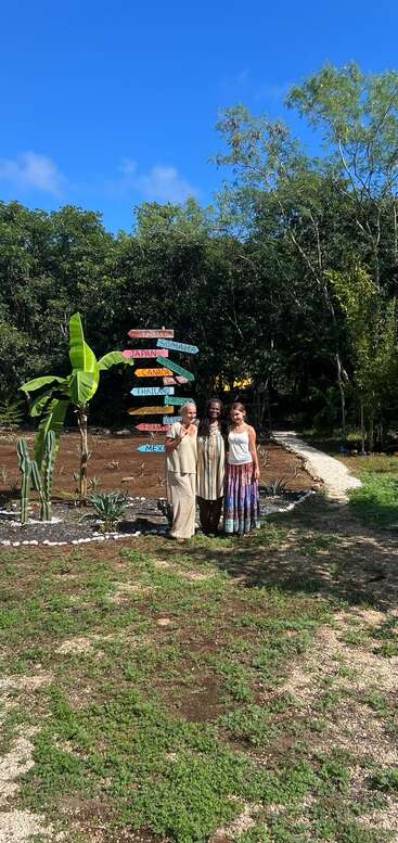 Drei Frauen stehen lächelnd neben einem bunten Wegweiser mit Ländernamen, umgeben von Grün, Bananenstauden, Kakteen und einem klaren blauen Himmel an einem sonnigen Tag.