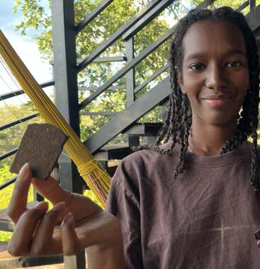 Eine lächelnde Person mit geflochtenen Haaren hält ein Stück braunes Material, möglicherweise Essen oder einen Stein, im Freien neben einer Treppe und einer gelben Hängematte, umgeben von Grünzeug.