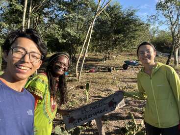 Tres amigos sonrientes posan juntos al aire libre en un soleado jardín rodeado de árboles y cactus, sosteniendo un cartel de madera hecho a mano en el que se lee "El Jardín, Mukammad, Brian."