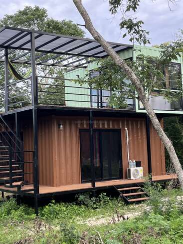 Esta imagen muestra una moderna casa de dos plantas construida con contenedores de transporte, rodeada de vegetación, con una terraza superior, escaleras, grandes ventanas y una hamaca.