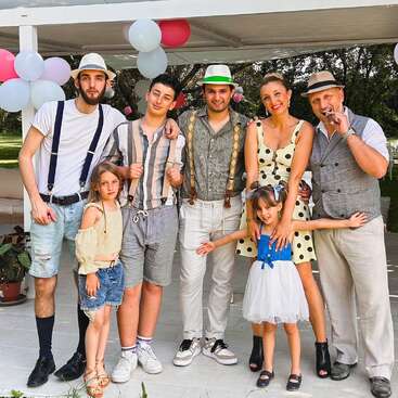 The image depicts a group of seven people, including two children, posing for a photo in front of a white pergola adorned with balloons, set against a lush green outdoor backdrop.
