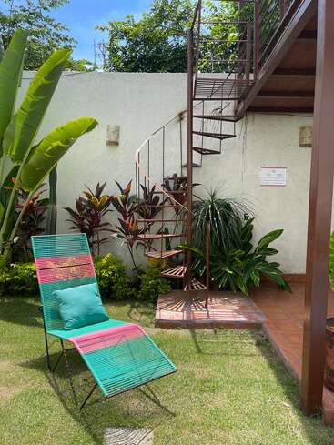 A colorful lounge chair sits on green grass near tropical plants. A spiral staircase leads up from a sunny patio against a white garden wall.