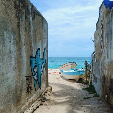 A sandy path between weathered concrete walls, one with blue shark graffiti, leads to a small blue boat and bright turquoise sea under a partly cloudy sky.
