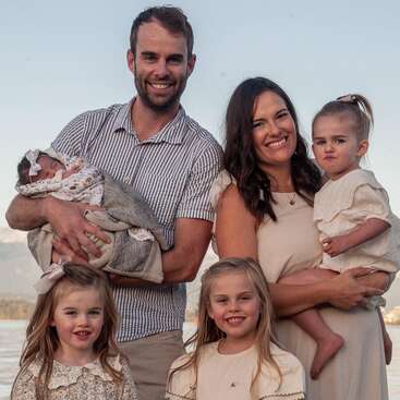 Une famille de six personnes pose au bord de l'eau, tout sourire. Les parents tiennent un bébé et un enfant en bas âge, tandis que deux petites filles se tiennent devant eux. Des montagnes se profilent en arrière-plan.