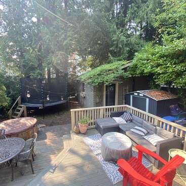 Une cour arrière chaleureuse comprend une terrasse en bois avec des meubles de détente, des chaises rouges, une table de jardin, un trampoline, un jacuzzi, un abri de jardin et un espace de rangement, le tout entouré de grands arbres.