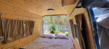 El interior de esta acogedora furgoneta camper cuenta con paneles de madera, una cómoda cama con ropa de cama de flores, ventanas con cortinas, suave luz natural y una apacible vista de la vegetación exterior.