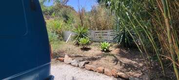 Una furgoneta azul está aparcada junto a un camino de grava bordeado de piedras. Exuberantes plantas verdes, bambú y una valla de madera blanca completan la soleada escena del jardín.