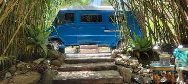 Una furgoneta azul de época está aparcada entre exuberante bambú y vegetación. Unos escalones de piedra conducen a la furgoneta, con asientos al aire libre y una pequeña mesa cerca. Un refugio tranquilo.