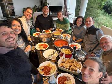 Un grupo de diez amigos se reúne alrededor de una mesa, sonriendo y compartiendo una comida a base de pasta y pan. El ambiente es cálido, alegre y acogedor.