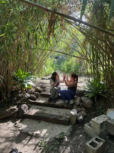 Dos personas se sientan frente a frente en unos rústicos escalones de madera rodeados de exuberante vegetación, jugando alegremente a las palmas bajo un arco de plantas altas.