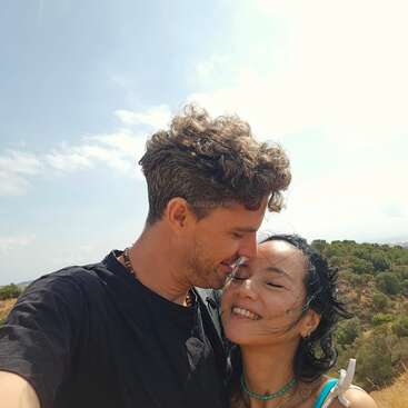 Un couple se tient à l'extérieur et s'embrasse affectueusement. L'homme appuie son front sur celui de la femme et tous deux sourient. Le ciel ensoleillé et les arbres verts constituent un arrière-plan naturel et serein.