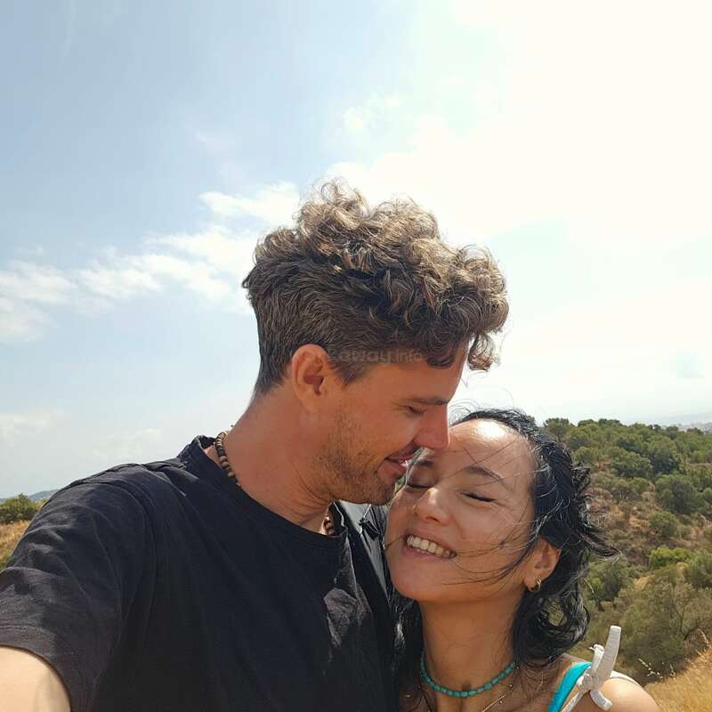 Una pareja se encuentra al aire libre, abrazándose cariñosamente. El hombre apoya la frente en la de la mujer y ambos sonríen. El cielo soleado y los árboles verdes proporcionan un fondo sereno y natural.