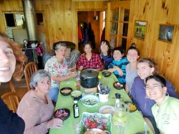 A group of nine people sit around a green table enjoying a meal together in a cozy, wooden dining room, smiling and sharing a joyful moment.