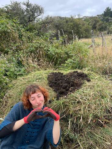 A woman with short, reddish hair and gardening gloves forms a heart shape with her hands. Behind her, soil is arranged in a heart on grassy ground.