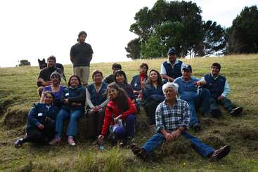 A group of people poses outdoors on a grassy hillside, with trees in the background. Some are sitting, others standing, and one dog is included.