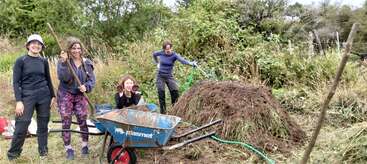 Four women work outdoors, smiling and posing with gardening tools by a wheelbarrow and a large compost pile, surrounded by green vegetation in a rural setting.