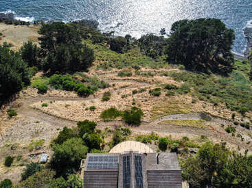 Uma vista aérea mostra uma casa com painéis solares, cercada por árvores e caminhos sinuosos, com vista para uma costa rochosa e um oceano cintilante sob a luz do sol.