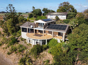 Esta imagem mostra uma casa moderna com grandes painéis solares, cercada por uma vegetação exuberante, um amplo deck de madeira e vistas panorâmicas do campo ao fundo.