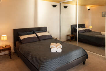 Um quarto aconchegante com uma cama de casal, roupa de cama cinza escuro, duas mesinhas de cabeceira com abajures, portas de armário espelhadas, paredes em tons neutros e toalhas cuidadosamente dobradas sobre a cama.