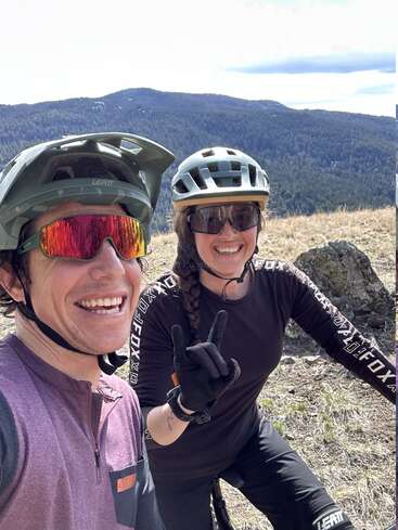 Two smiling cyclists wearing helmets and sunglasses take a selfie in a sunny mountain landscape. One makes the “rock on” hand sign. Both look happy.