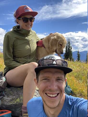 A smiling man takes a selfie outdoors with a woman and a golden retriever dog. They sit on a rock amid grassy fields, mountains, and blue sky.