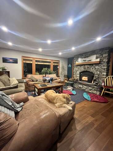 A cozy living room with a stone fireplace, dog lounging on a couch, another dog by coffee table, soft lighting, wooden floors, and comfortable furniture.