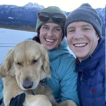 A happy couple and their golden retriever pose for a selfie outdoors, bundled in winter jackets and hats, with snowy mountains in the background. Smiling, joyful moment.