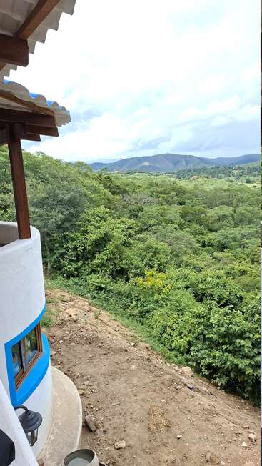 A white curved building with blue trimming overlooks a lush green forest. Dirt ground in front, distant mountains under a cloudy sky complete the scenic view.