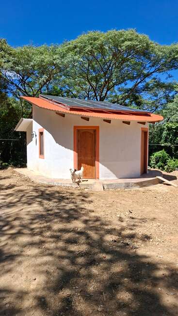 A small white house with an orange roof trim, solar panels, a wooden door, a dog standing outside, surrounded by trees and blue sky.
