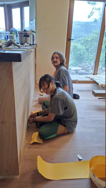Two people sit on a wooden floor, sanding a wooden surface. One smiles playfully at the camera, while sunlight streams through large windows, revealing a scenic landscape.