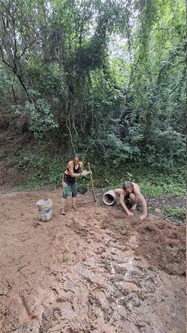 Two people are working outdoors in a muddy clearing near a forest. One is using a stick, the other crouches, mixing or spreading mud and natural fibers.